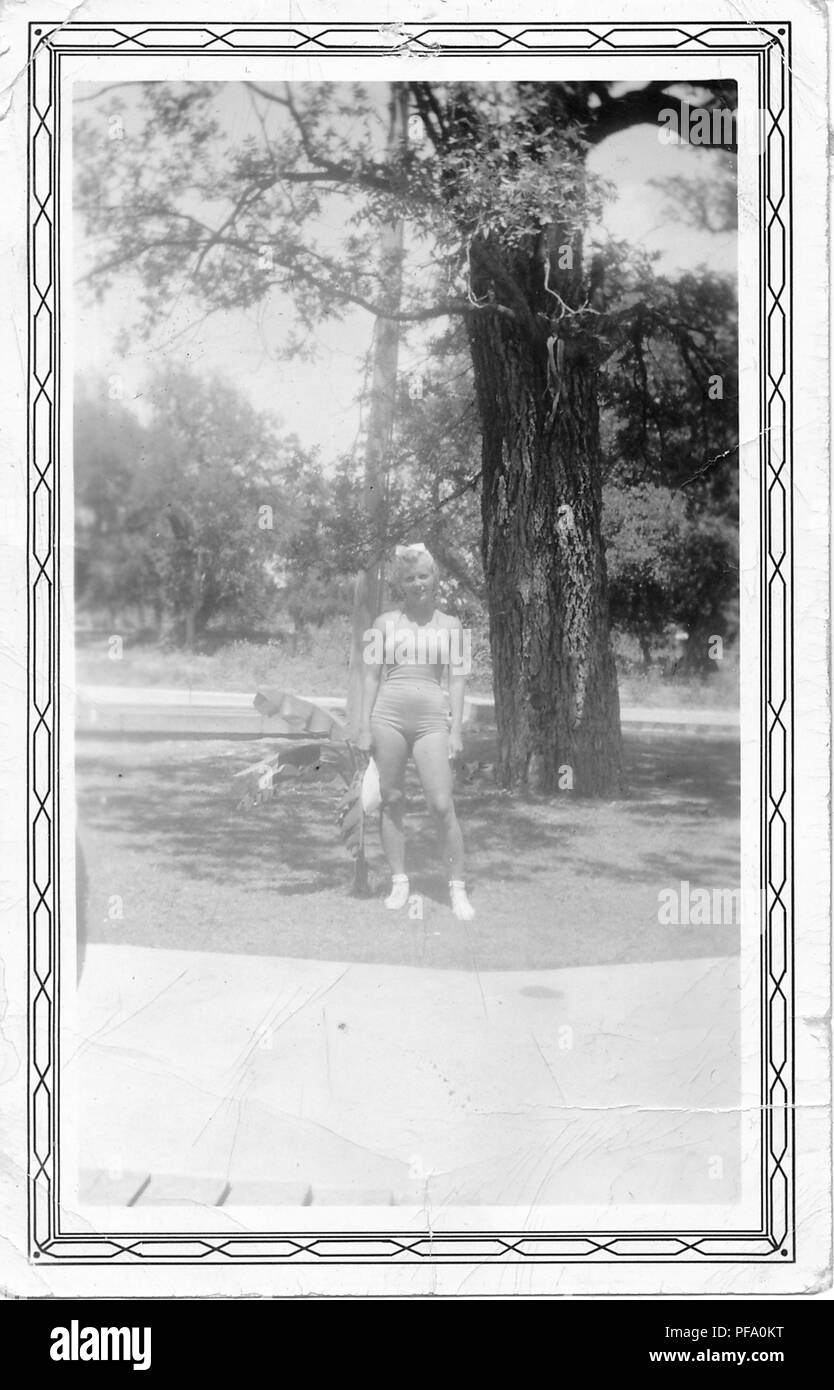 Photographie noir et blanc, montrant une blonde aux cheveux bouclés, femme, en vue de pleine longueur, extérieur, face à la caméra, portant une lumière colorée, une pièce maillot de bain, et la tenue d'un pare-soleil, avec un petit palmier et grand arbre visible à l'arrière-plan, probablement photographié en Ohio dans la décennie suivant la Seconde Guerre mondiale, 1950. () Banque D'Images