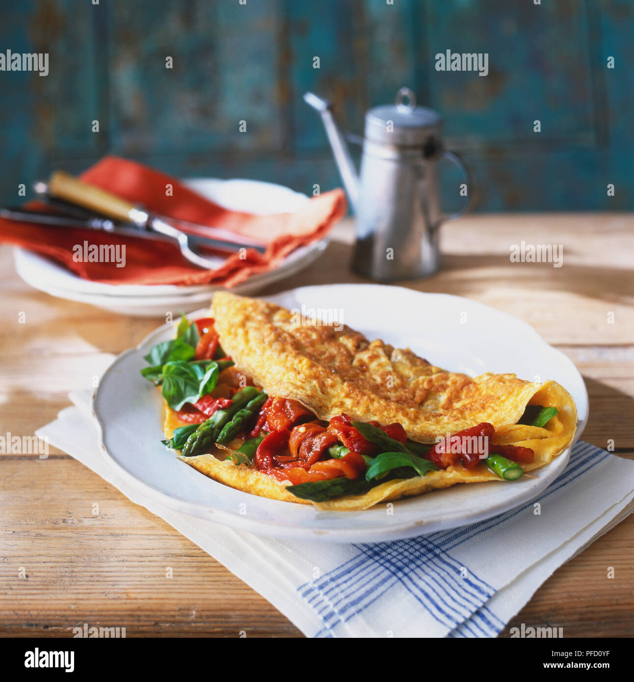 Omelette, farci aux asperges vertes, tranches de poivron rouge et l'ensemble de feuilles de basilic, servi sur une plaque blanche, sur la table de bois rustique, avec des couverts de plaques empilées sur une serviette rouge, tin pot avec buse longue en arrière-plan Banque D'Images