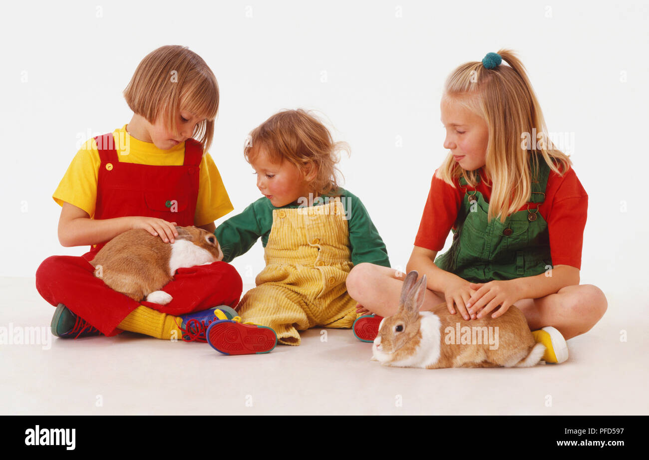 Trois blondes assise sur le sol en jouant avec deux lapins (Oryctolagus cuniculus), vue de face. Banque D'Images