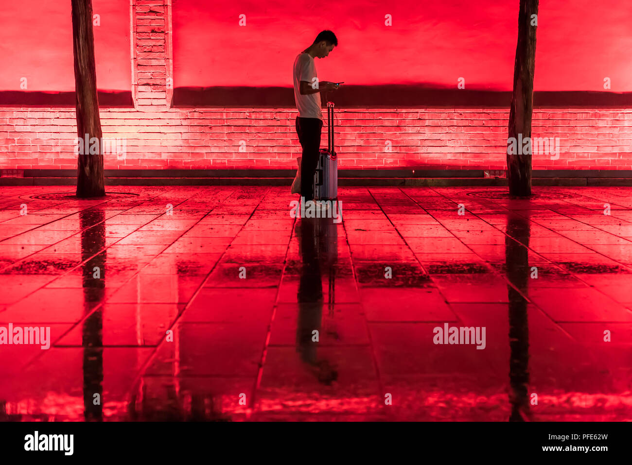 Xi'an, province du Shaanxi, Chine - Aug 9, 2018 : tourisme chinois avec sa valise à la recherche de son smartphone de nuit sous la pluie avec l'environnement de lumière rouge dans la rue. Banque D'Images