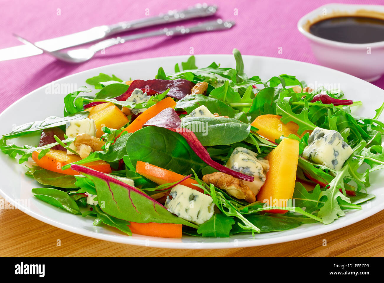 Kaki, fromage bleu, roquette, jeunes épinards, laitue et noix délicieuse salade sur assiette blanche sur tapis de table avec une fourchette, couteau, close-up, macro Banque D'Images