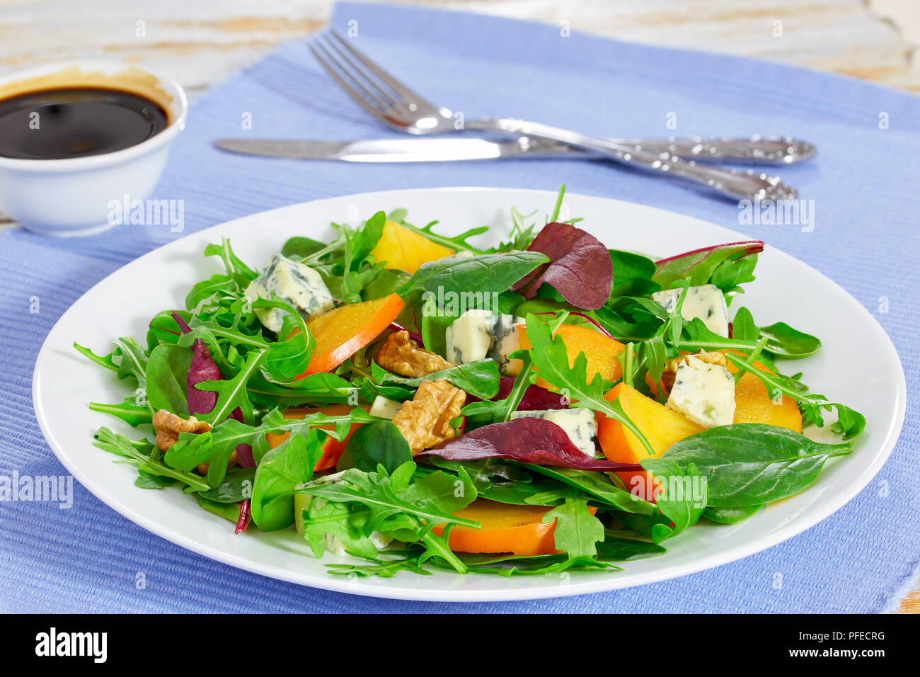 Kaki, fromage bleu, roquette, jeunes épinards, laitue et noix délicieuse salade sur assiette blanche sur tapis de table avec la fourchette, le couteau et le vinaigre dres Banque D'Images