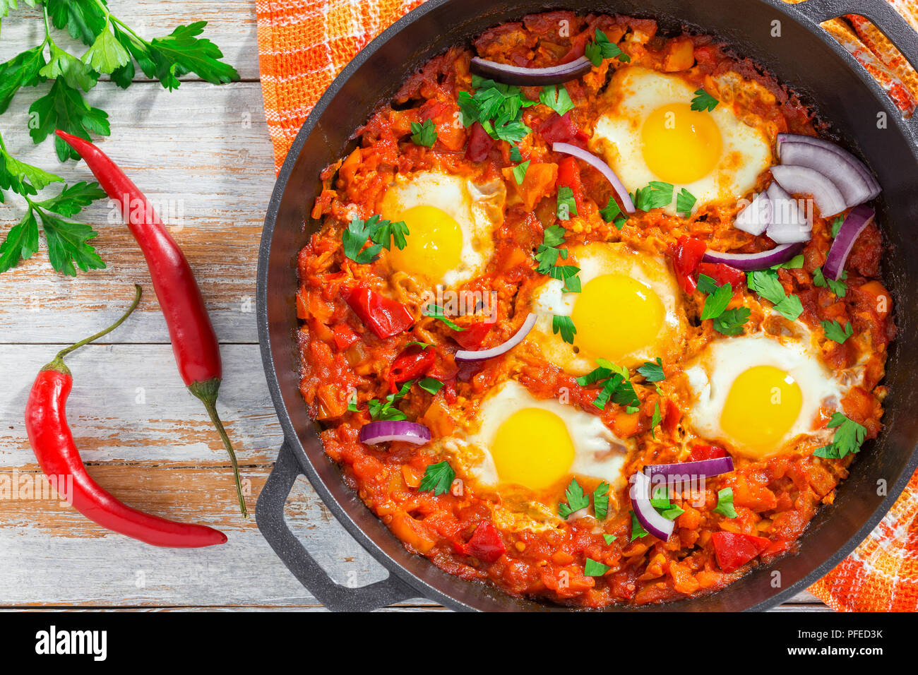 Petit-déjeuner sain shakshuka - œufs frits, oignon, poivron, tomate, piment et épices dans poêle de fer avec une serviette de cuisine, le persil sur planche en bois blanc Banque D'Images