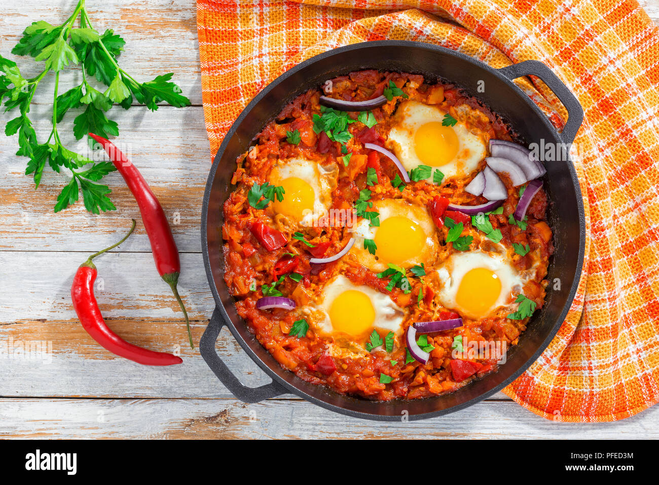 Petit-déjeuner sain shakshuka - œufs frits, oignon, poivron, tomate, piment et épices dans poêle de fer avec une serviette de cuisine, le persil sur planche en bois blanc Banque D'Images