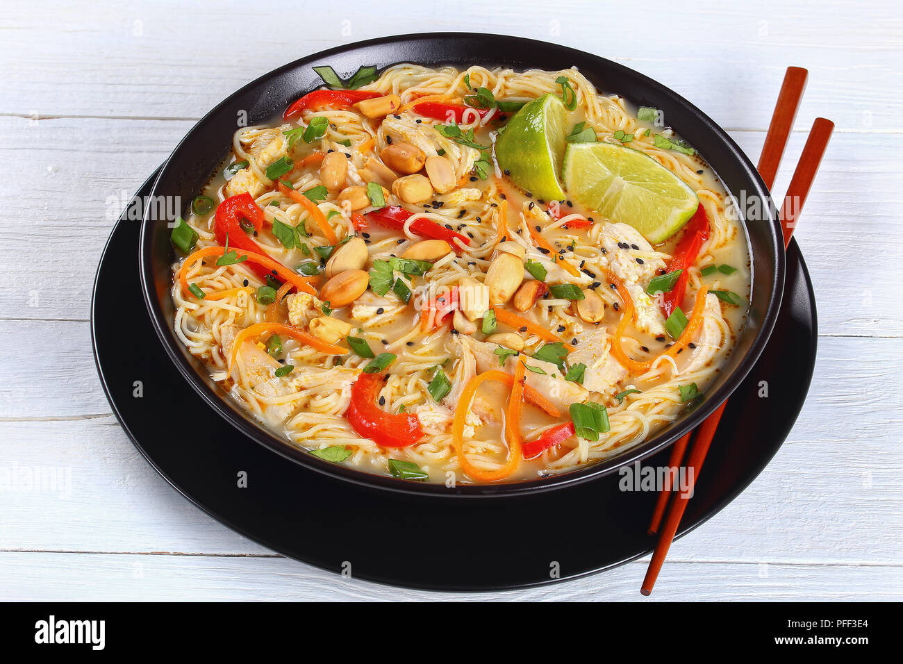 Soupe Poulet et nouilles thaï saupoudré de coriandre, de sésame et d'arachide dans un bol avec des baguettes noires. l'oignon vert, la moitié de poivron rouge et g Banque D'Images