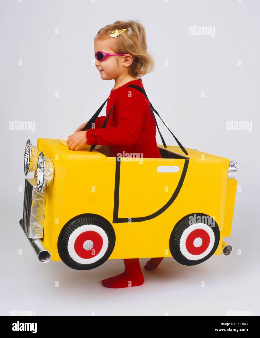 Petite fille blonde portant des vêtements et des lunettes de soleil, les sangles autour de l'épaule, debout en jaune carton car, sous les jambes, bouclier, grill et lumières enveloppées dans du papier d'aluminium, papier debout sur la plaque de bonnet Banque D'Images