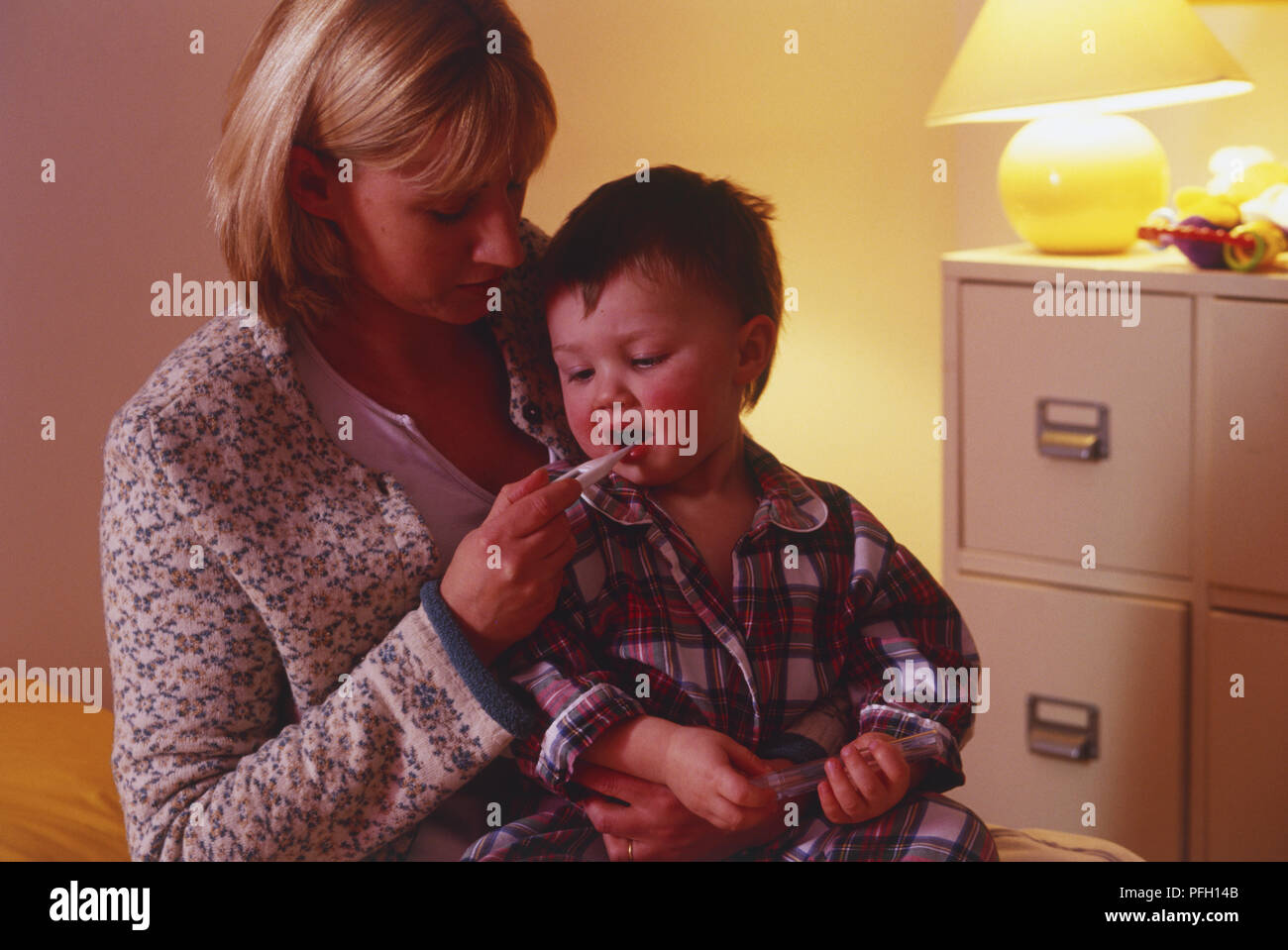Femme assise avec tout-petit garçon sur ses genoux, holding digital thermomètre dans la bouche du garçon. Banque D'Images
