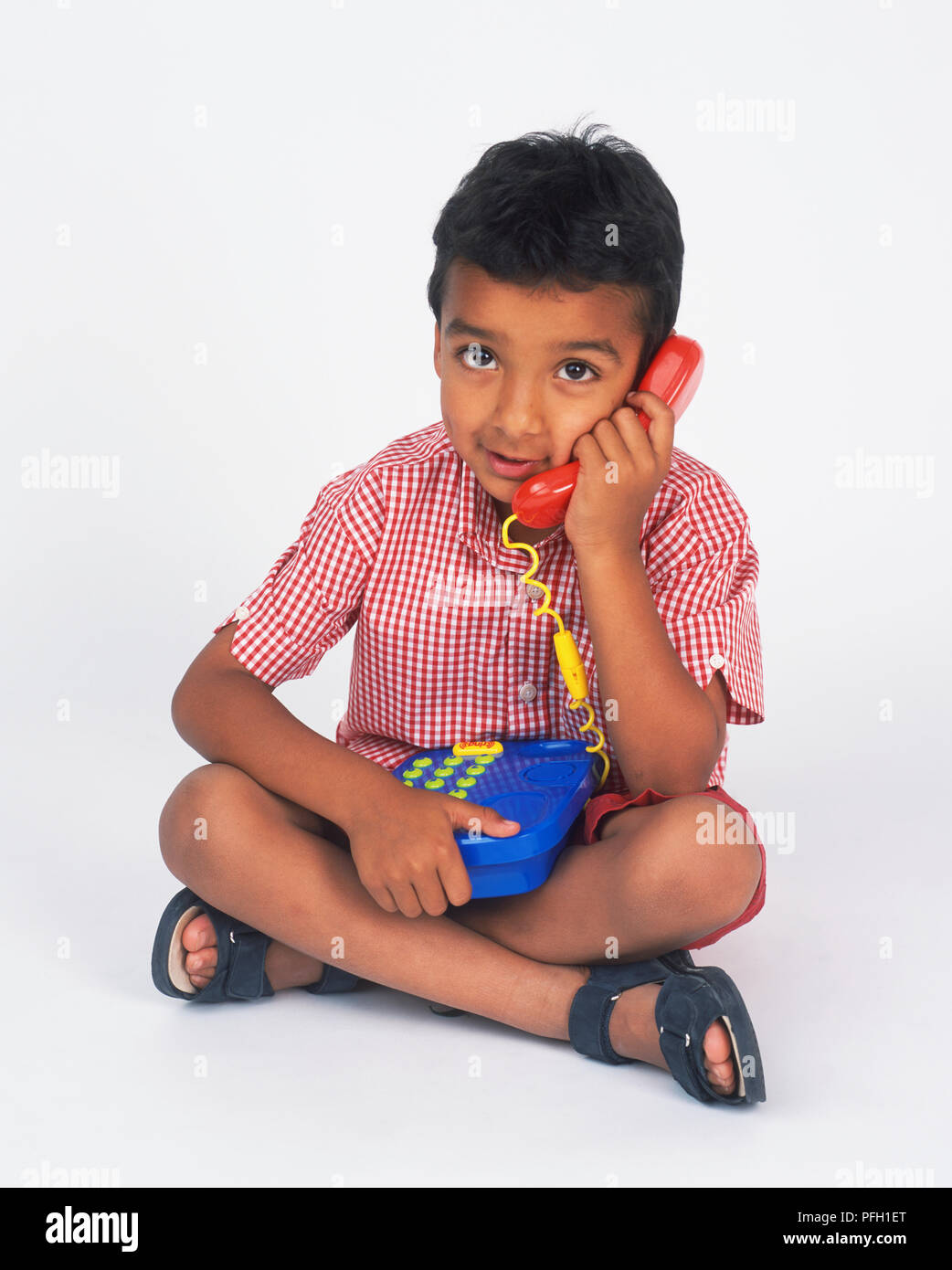 Boy sitting cross legged, holding toy téléphone sur ses genoux et le récepteur à son oreille Banque D'Images