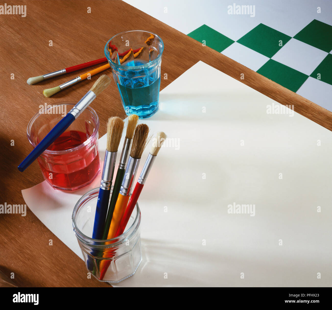 Assortiment de pinceaux dans un bocal en verre à côté de verres de couleur rouge et bleu de l'eau, feuille de papier sur la table. Banque D'Images