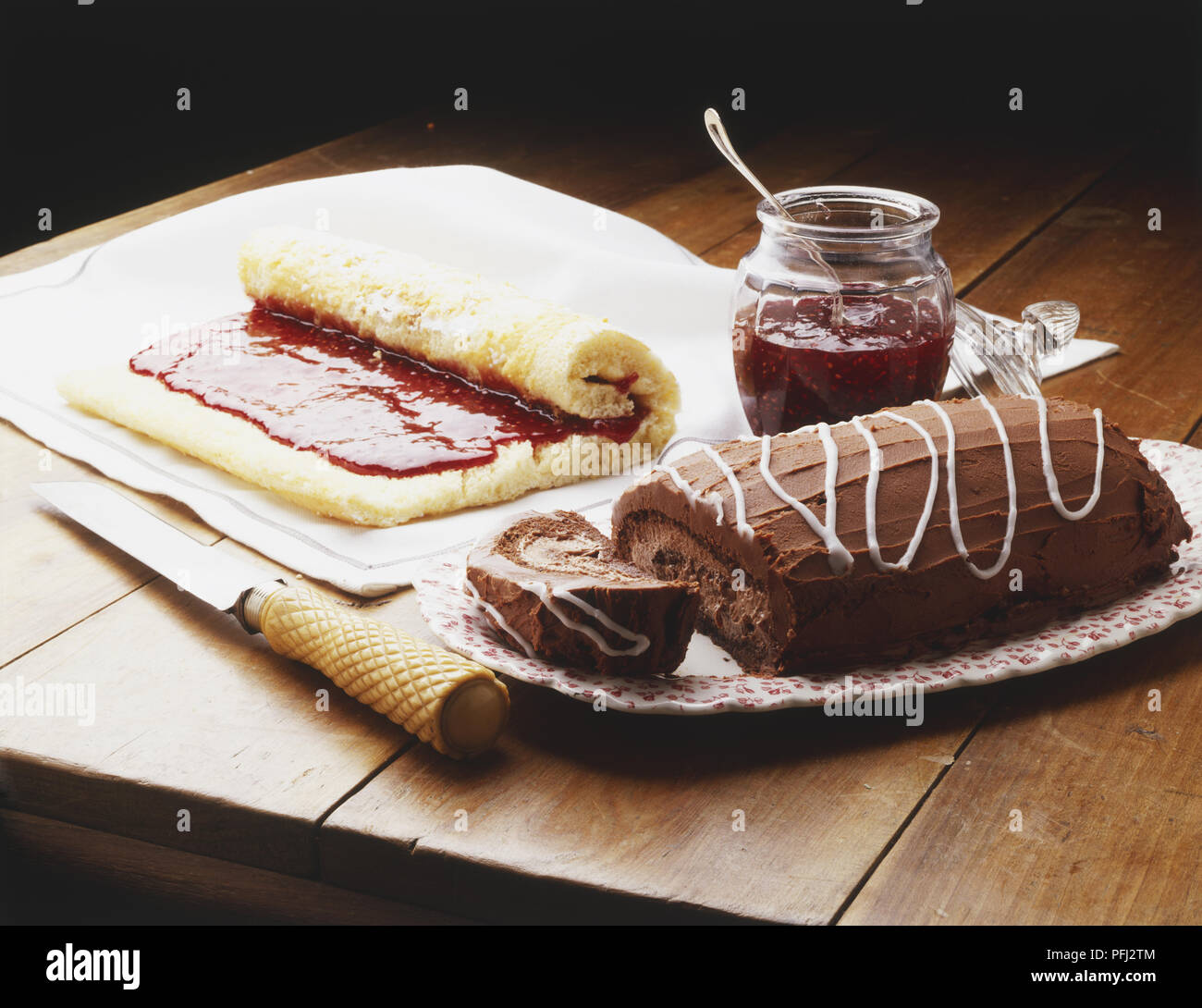 En partie enroulé jelly roll montrant couche de gelée, à côté de pot de gelée et une crème moka rouler sur un plat de service, sur une table en bois Banque D'Images