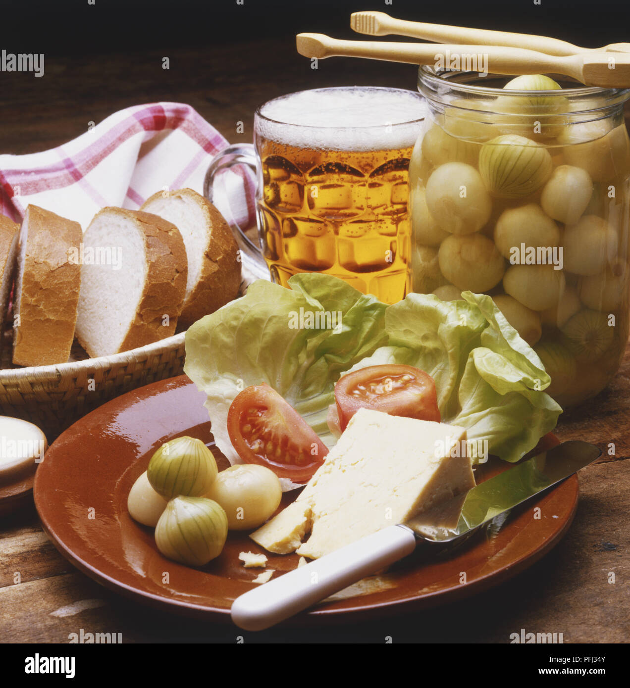 Fromage, salade et oignons marinés sur une plaque, panier contenant des tranches de pain blanc, verre de bière et pot d'oignons confits Banque D'Images