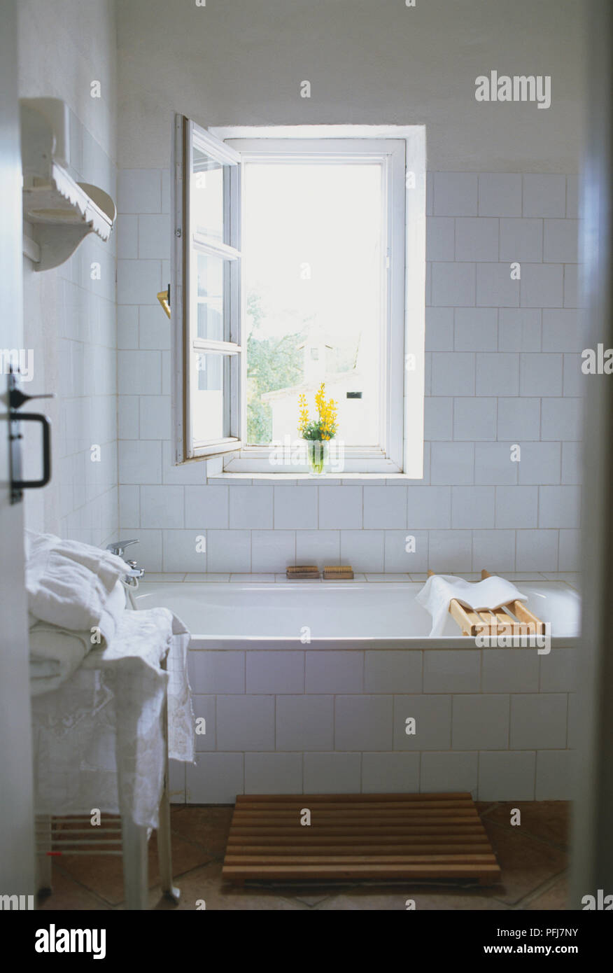 Salle de bains avec murs blancs et bassin rectangulaire, table avec les  serviettes blanches, carrelage en terre cuite avec un tapis de bain,  fenêtre ouverte, fleurs jaunes sur rebord de fenêtre Photo