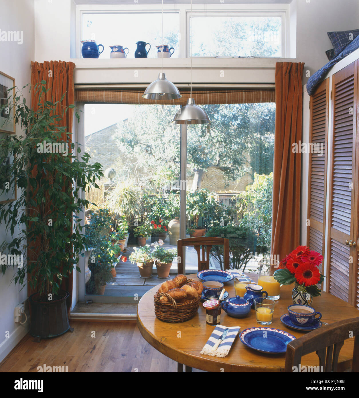 Table de cuisine ronde prévue pour le petit déjeuner, de grandes portes en verre en arrière-plan une vue sur patio en bois rempli de plantes en pot. Banque D'Images