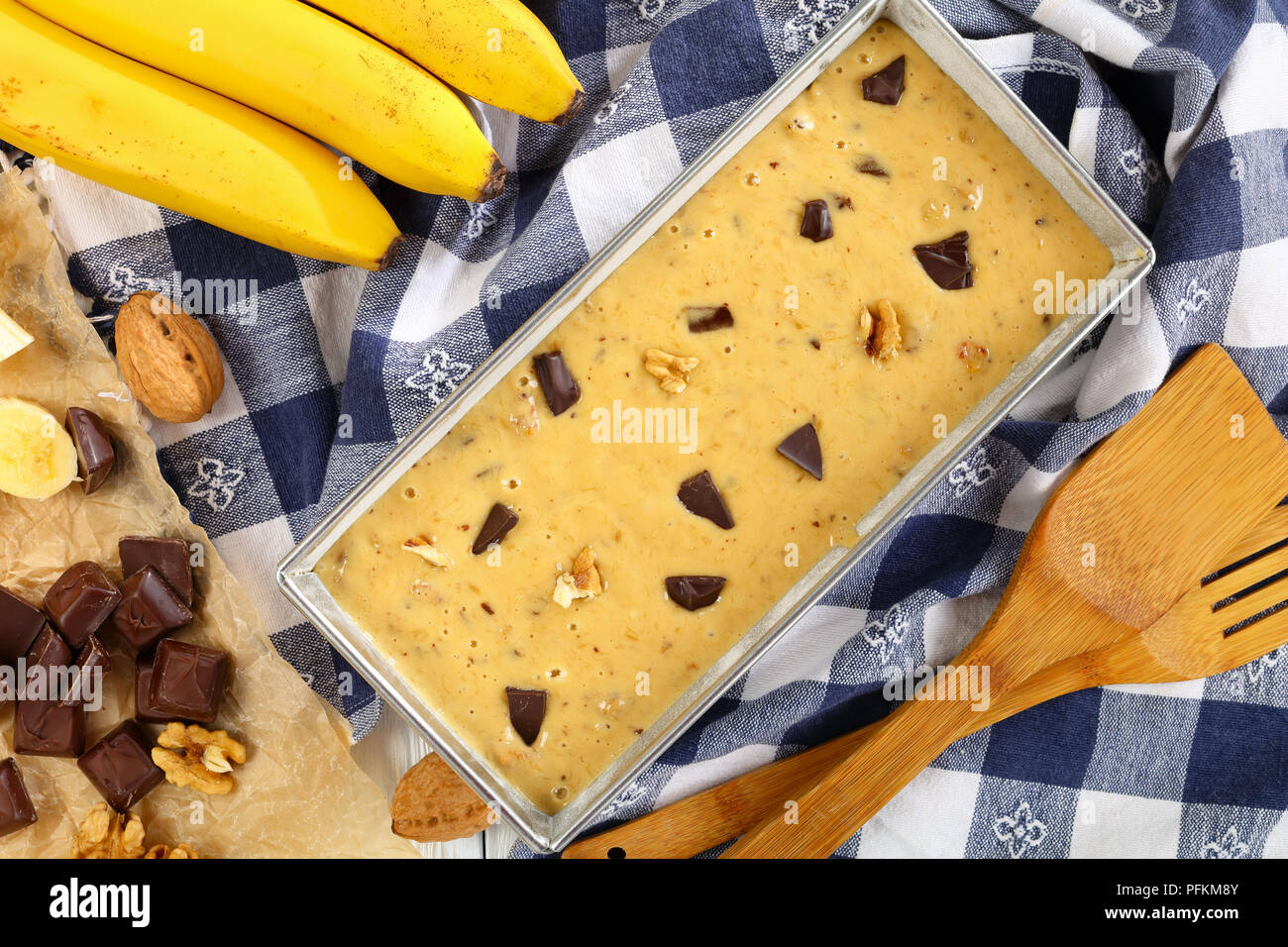 Pain aux bananes et aux noix brutes et de morceaux de chocolat dans un moule à pain sur table en bois avec des serviettes de cuisine, spatules et les ingrédients à l'arrière-plan, vue depuis Banque D'Images
