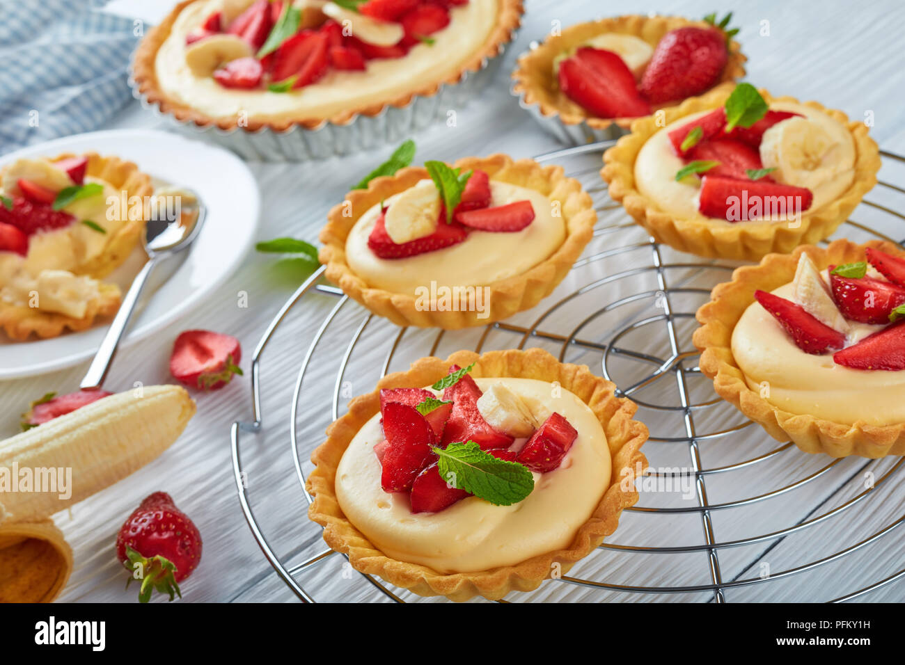 De délicieuses tartelettes aux fraises, des tranches de banane chargé avec crème pâtissière sur une grille, et sur une plaque blanche avec une cuillère, vue de dessus, à proximité- Banque D'Images