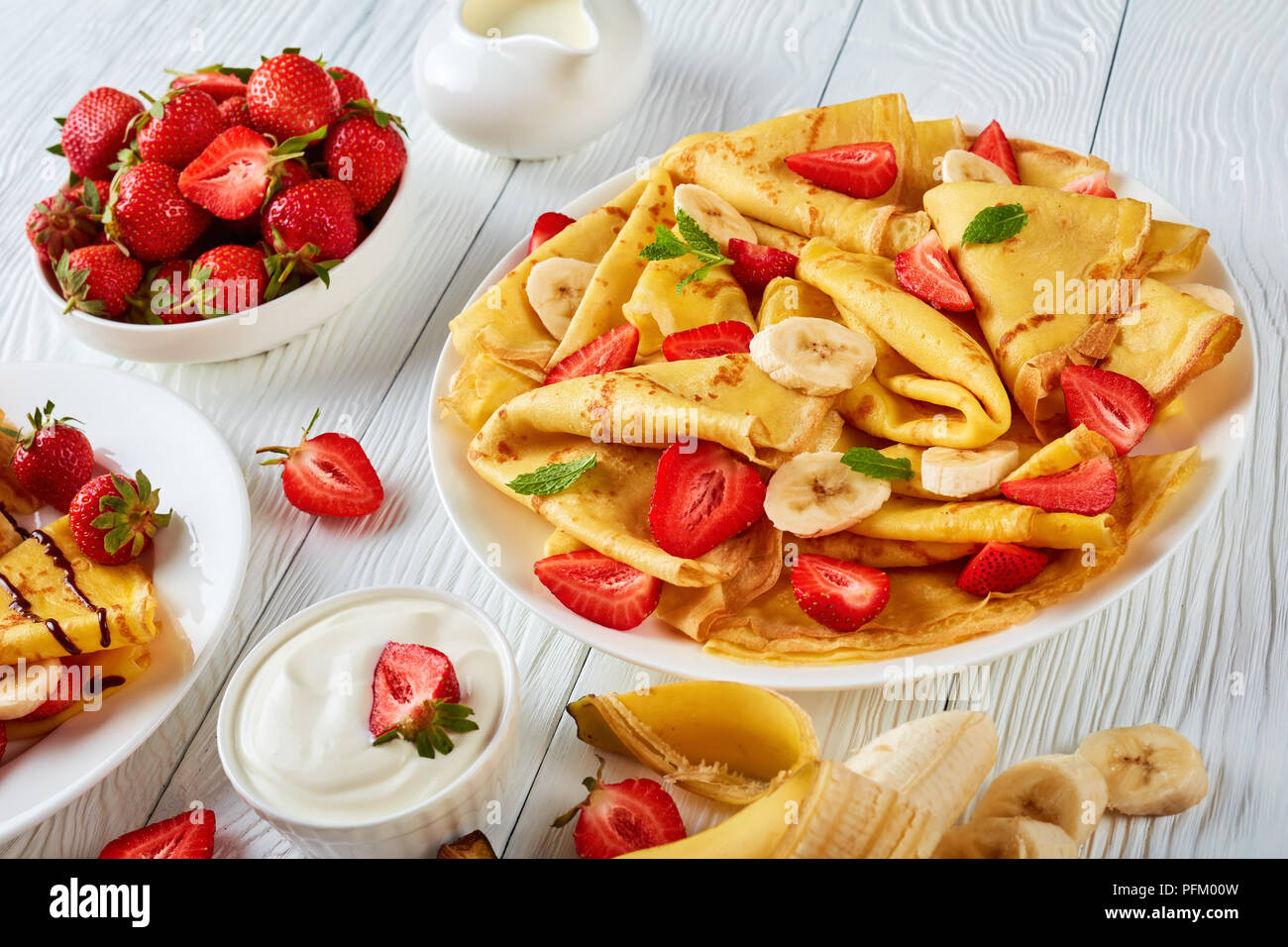 Crêpes minces ou des crêpes sur une plaque avec des fraises, des tranches de banane et de feuilles de menthe, la crème et le bol avec des fraises fraîches à l'arrière-plan, ho Banque D'Images