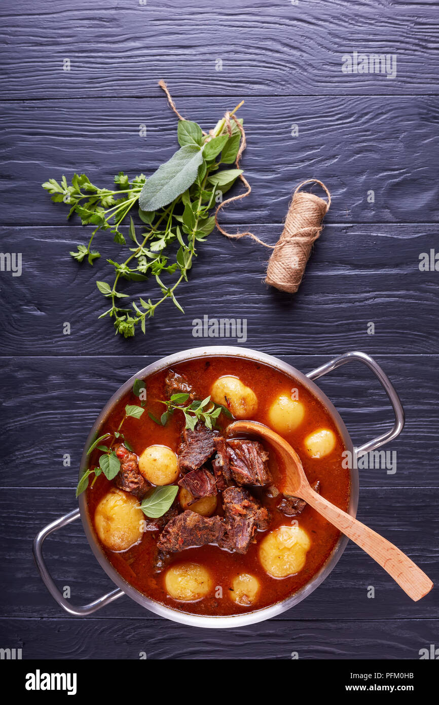 Délicieux Ragoût de boeuf avec des cubes de viande tendre, ensemble de nouvelles pommes de terre, carottes et herbes dans un chaudron sur une table en bois noir avec son bouquet aromatique de Banque D'Images
