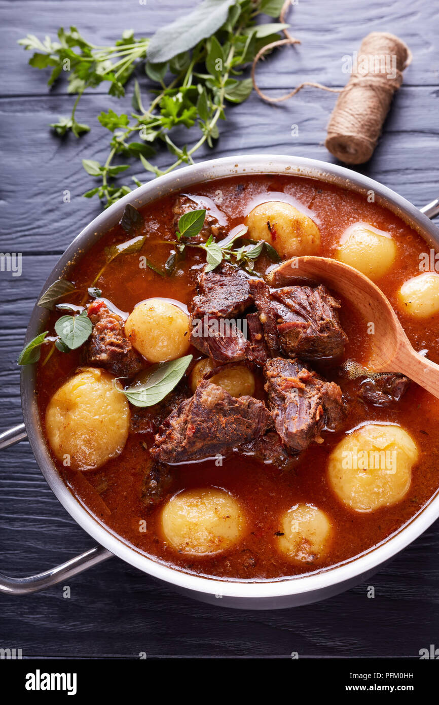 Ragoût de boeuf avec des cubes de viande tendre, ensemble de nouvelles pommes de terre, carottes et herbes dans un chaudron sur une table en bois noir avec bouquet d'herbes aromatiques, vertic Banque D'Images