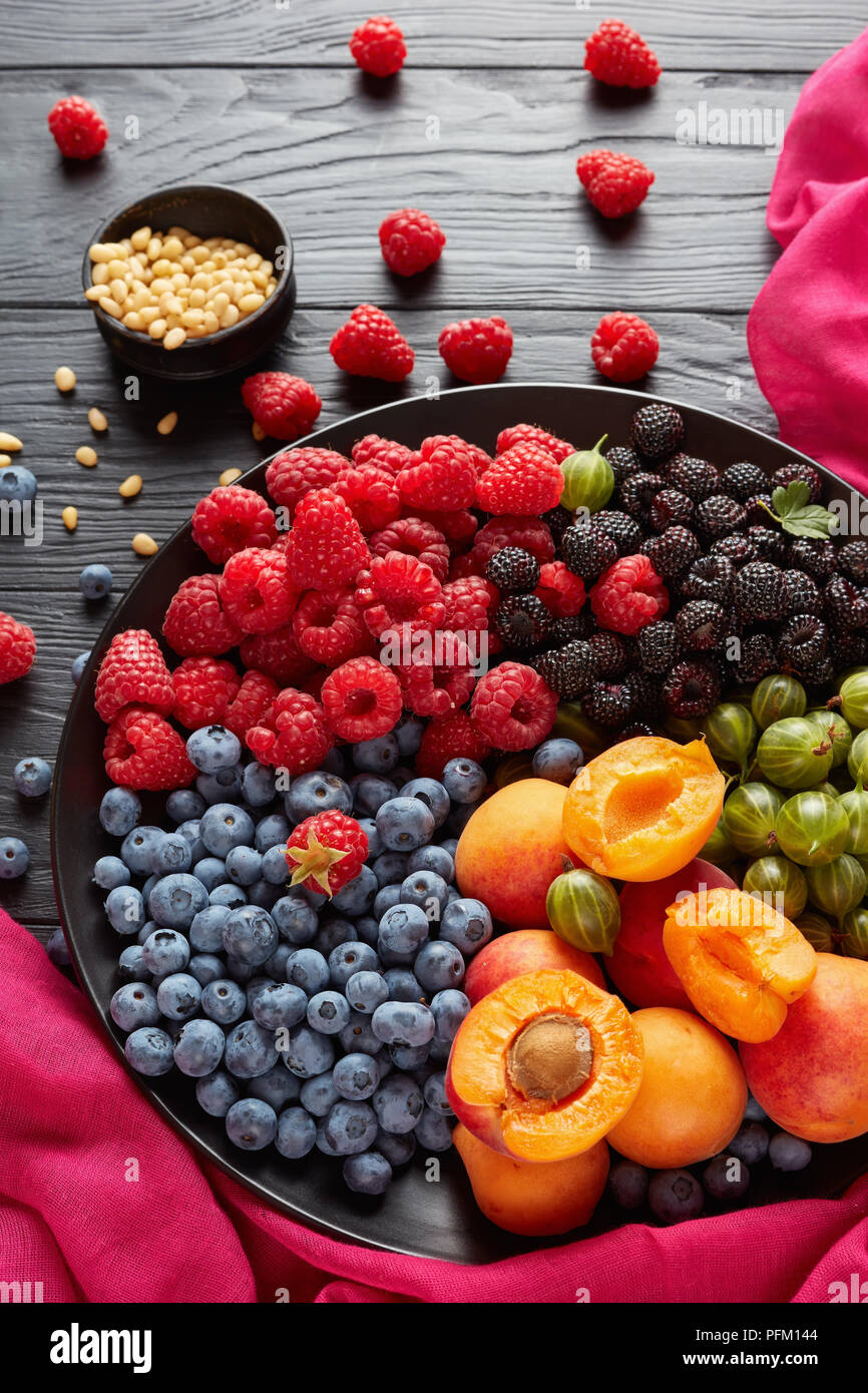 Close-up de l'assiette de fruits et baies avec des produits bio frais de groseille, framboise rouge et noire, de bleuet, de tranches d'abricots sur une plaque noire sur un sol en bois t Banque D'Images