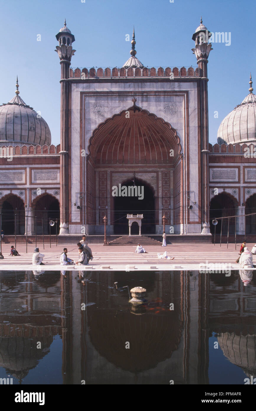 L'Inde, Delhi, la grande mosquée Jami Masjid, construit en 1656 par l'empereur Shah Jahan, la plus grande mosquée de la congrégation en Asie, grande entrée, deux dômes, les gens devant elle, reflétant dans l'eau en premier plan. Banque D'Images
