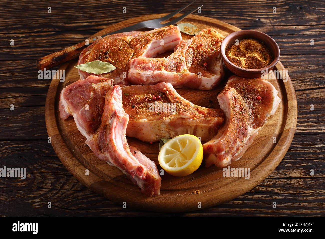 Close-up de côtelettes de porc non cuit sur l'os assaisonnée d'épices, de poivre et de laurier sur une planche en bois avec fourchette à découper sur ta rustique en bois Banque D'Images