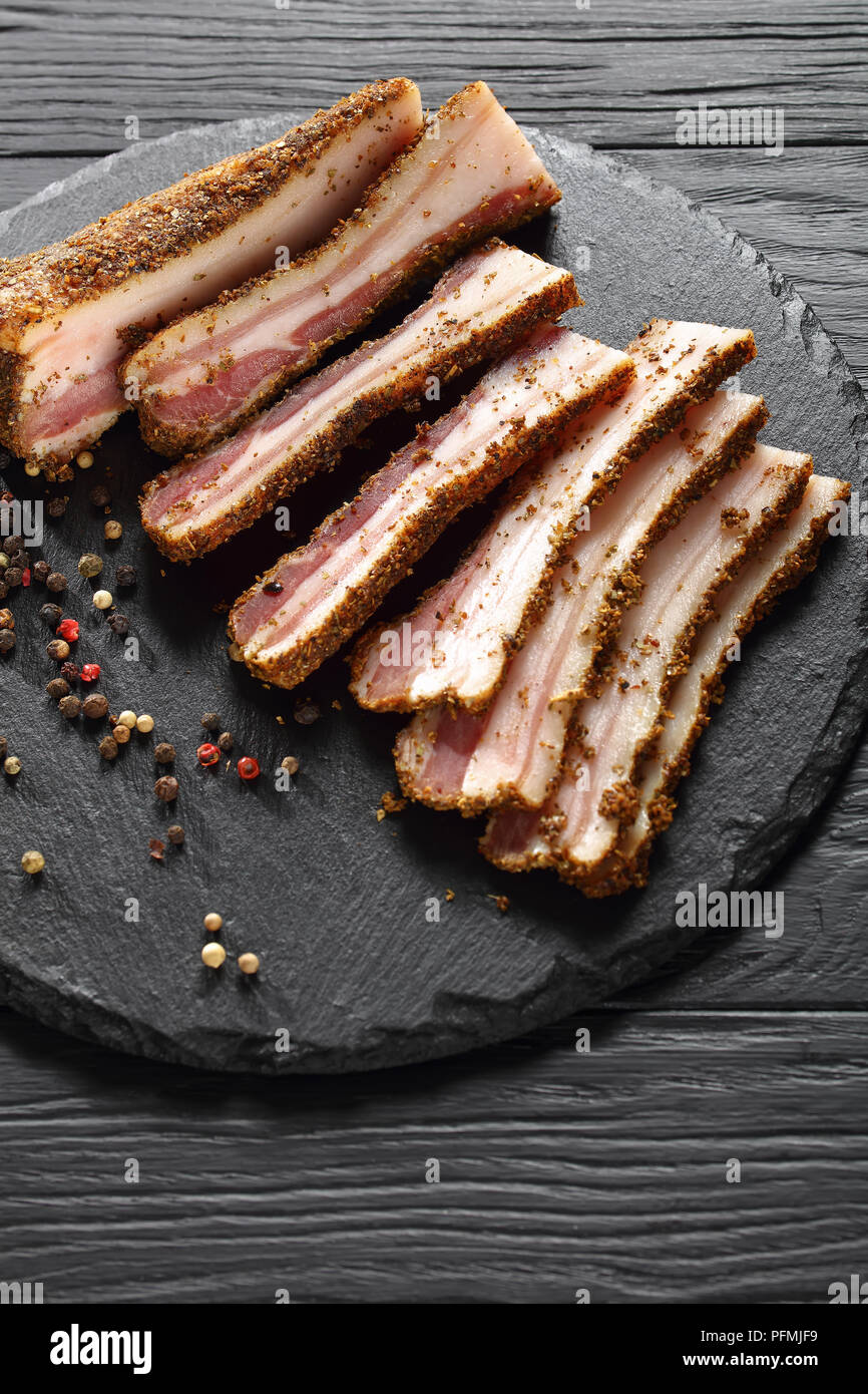 Speck fumé épicé délicieux coupé en tranches sur la plaque en pierre ronde noire sur la table en bois, vue verticale d'en haut Banque D'Images