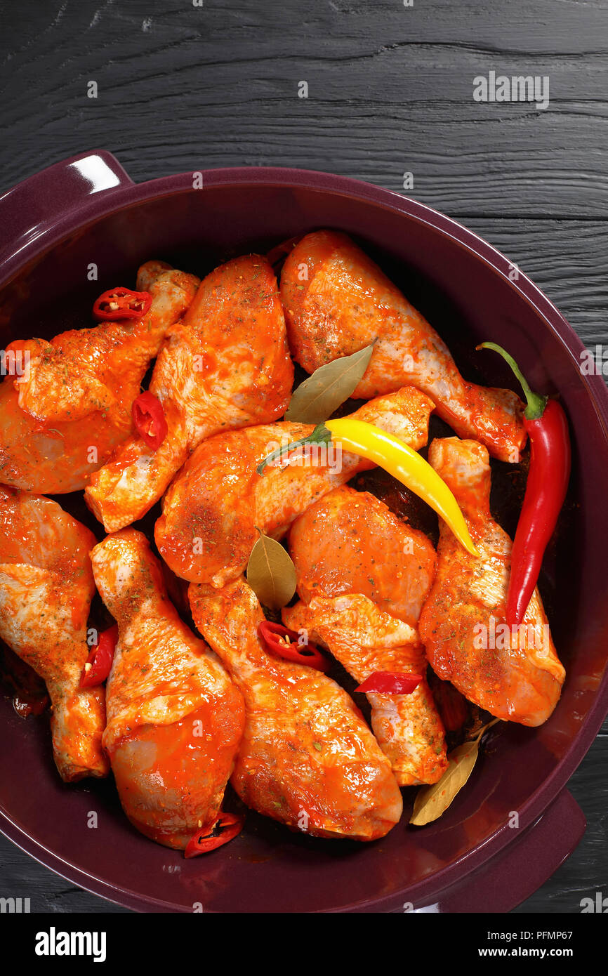 Pilons de poulet cru mariné frotté avec la spice, morceaux de piment, les feuilles de laurier prêt à cuire dans un plat, vue verticale de abov Banque D'Images