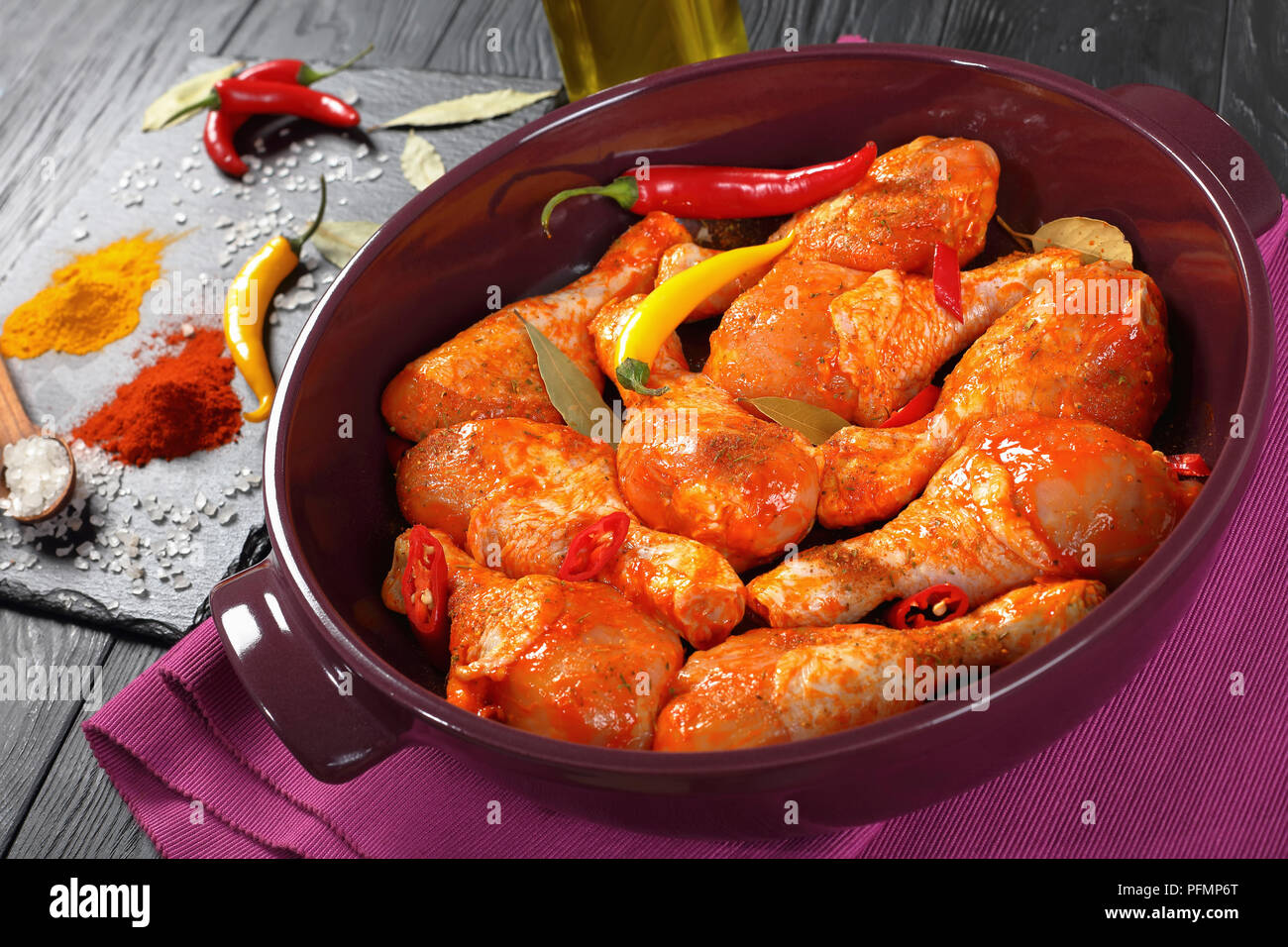 Pilons de poulet cru mariné saupoudré d'épices, morceaux de piment, les feuilles de laurier prêt à cuire dans un plat, les ingrédients sur une pierre sur une carte Banque D'Images