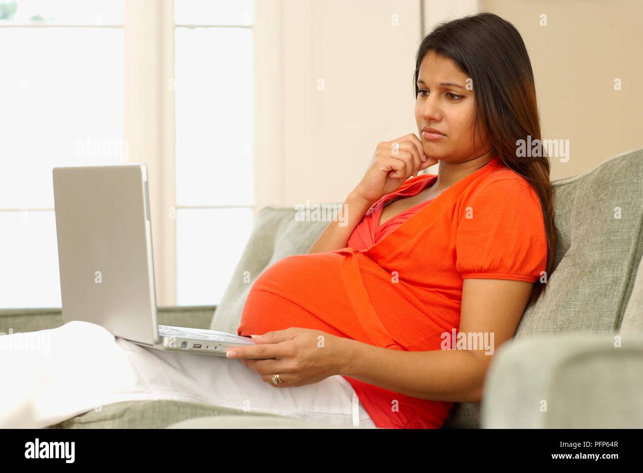 Pregnant woman sitting on sofa looking at laptop écran, side view Banque D'Images