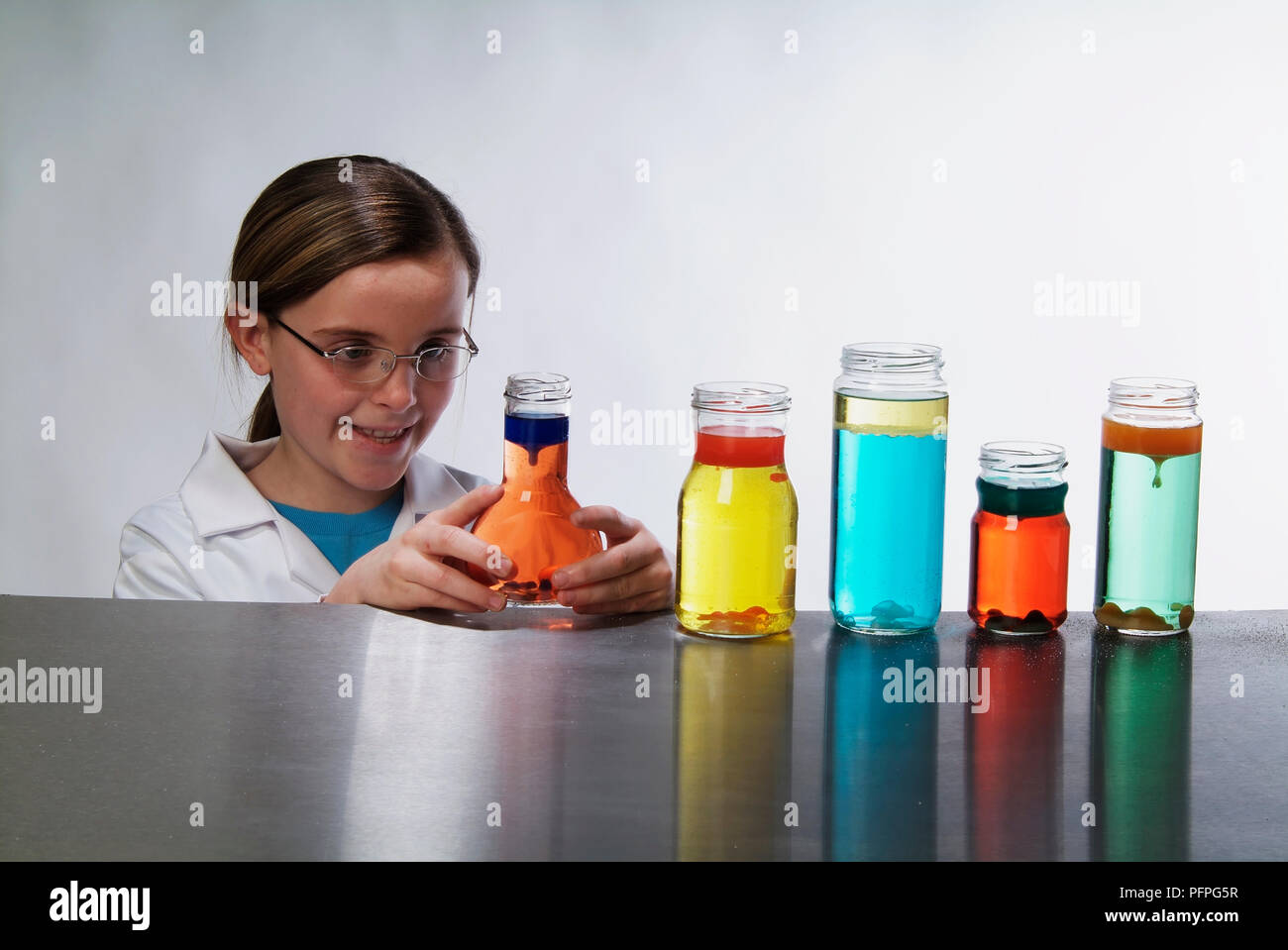 Pré-adolescente in lab coat, souriant, l'examen de l'un des cinq petites bouteilles contenant des liquides colorés sur la table Banque D'Images