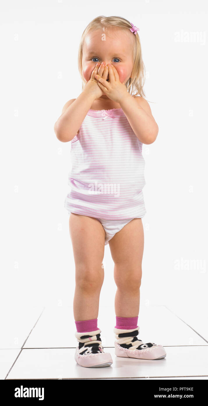 Girl wearing vest, pantalon et chaussons, couvrant son visage avec ses mains, 20 mois Banque D'Images