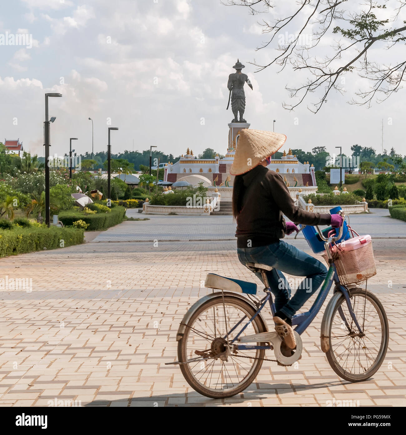 Une femme laotienne conduit une bicyclette dans le parc Chao Anouvong avec la grande statue du roi Chao Anouvong en arrière-plan, à Vientiane, Laos Banque D'Images
