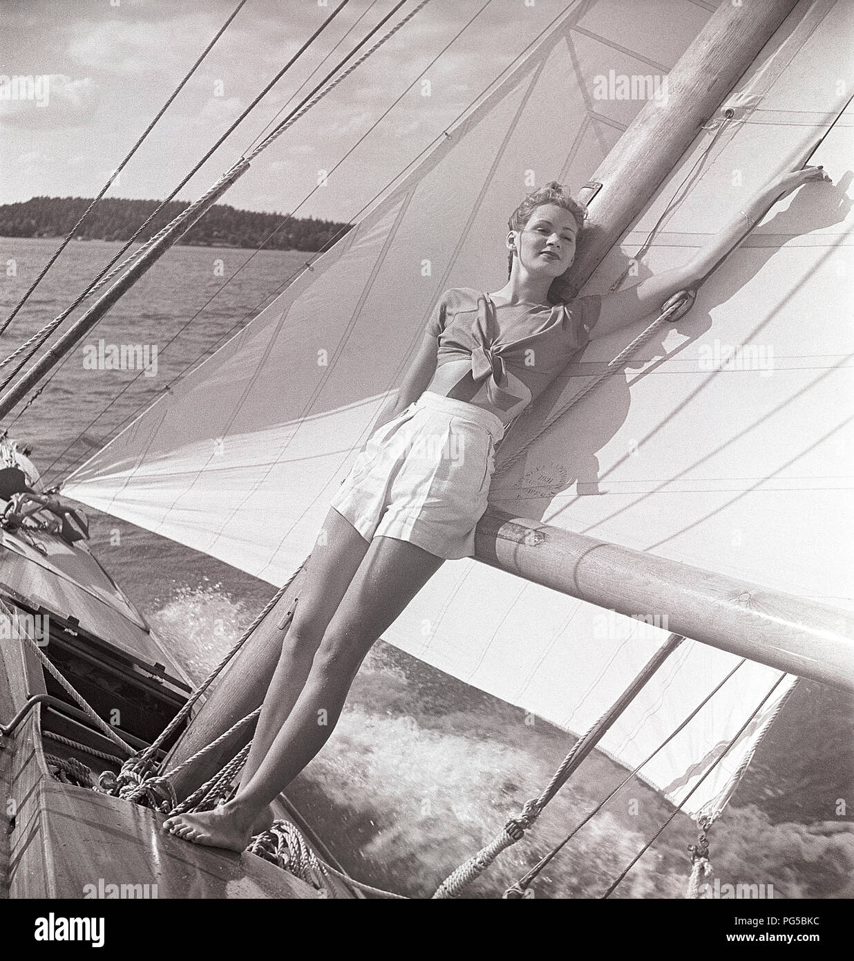 1940 Bateau à voile. Une jeune femme est à bord d'un bateau à voile et debout sur le pont et se penchant sur le mât et la voile quand il cruises avant avec le vent dans les voiles. Elle est vêtue d'une façon typique des années 40 avec une chemise et un short. La Suède 1946 Photo Kristoffersson ref AC101-2 Banque D'Images