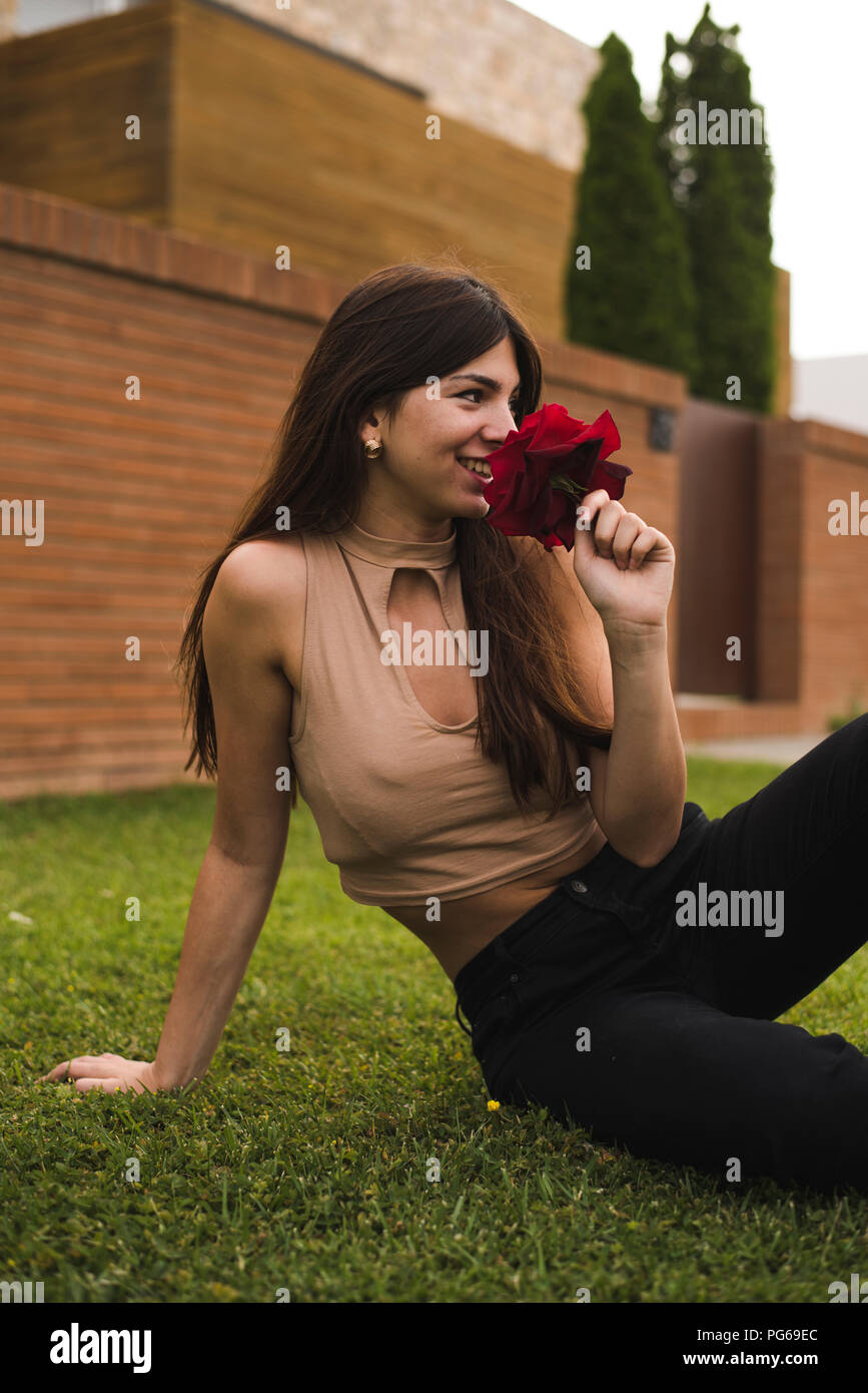 Smiling young woman sitting on lawn smelling red rose Banque D'Images