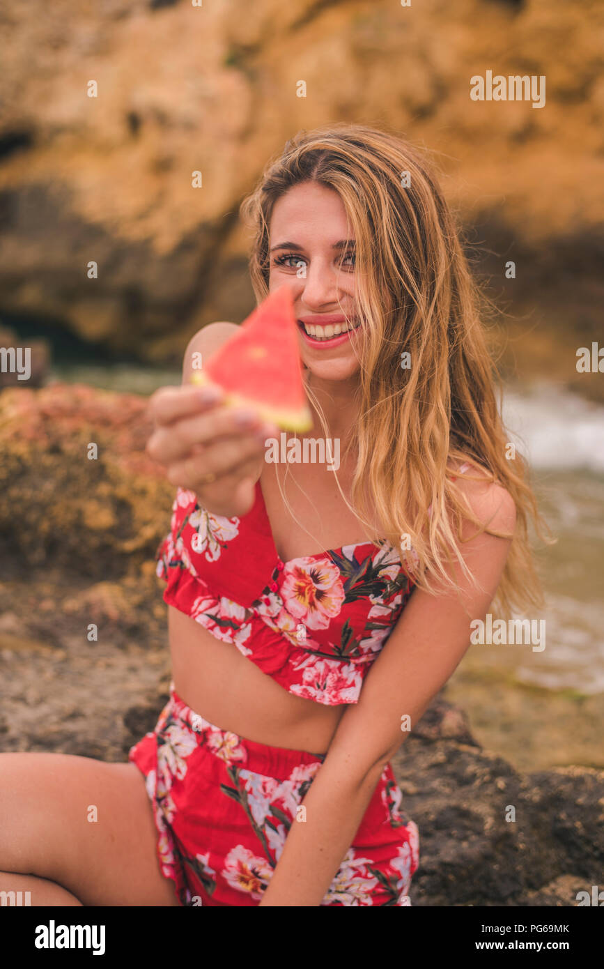 Portrait of happy young woman on rocks at the sea holding watermelon slice Banque D'Images