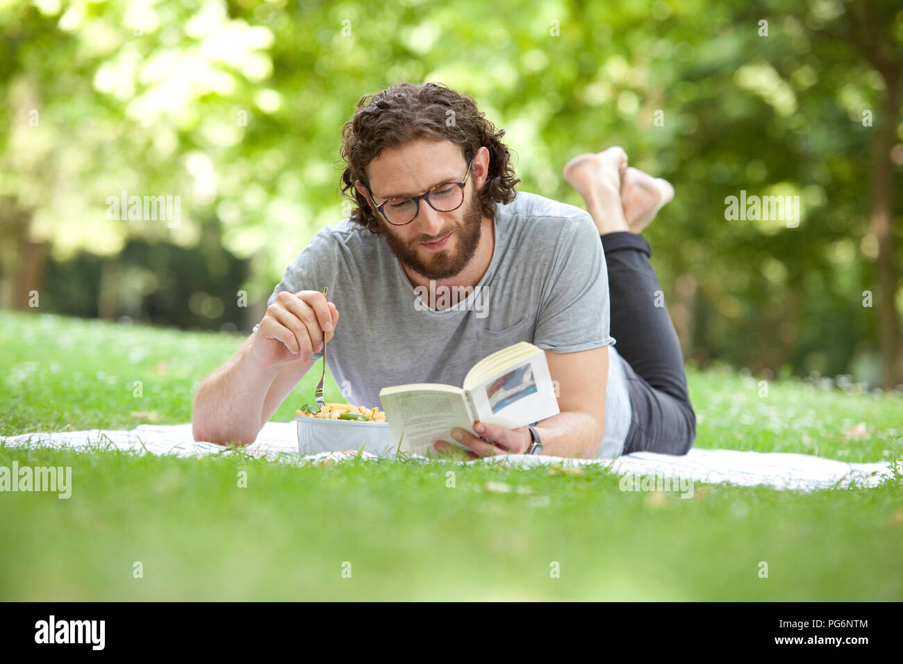 Man reading book on blanket in a park en mangeant une salade de nouilles Banque D'Images