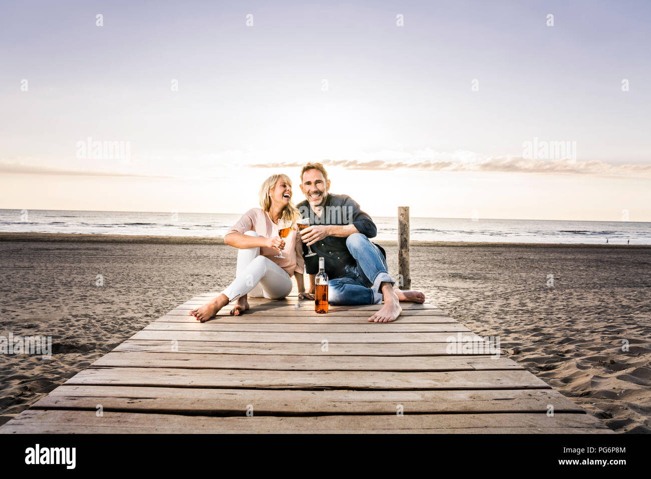 Couple heureux avec des verres à vin assis sur la demande sur la plage au coucher du soleil Banque D'Images