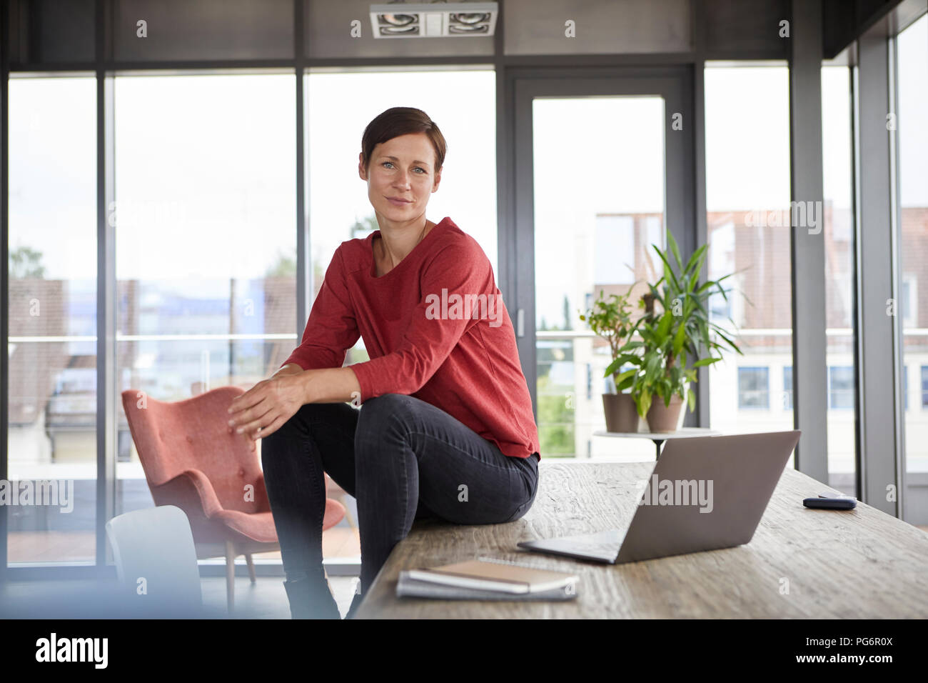 Portrait of smiling woman sitting on table at home with laptop Banque D'Images