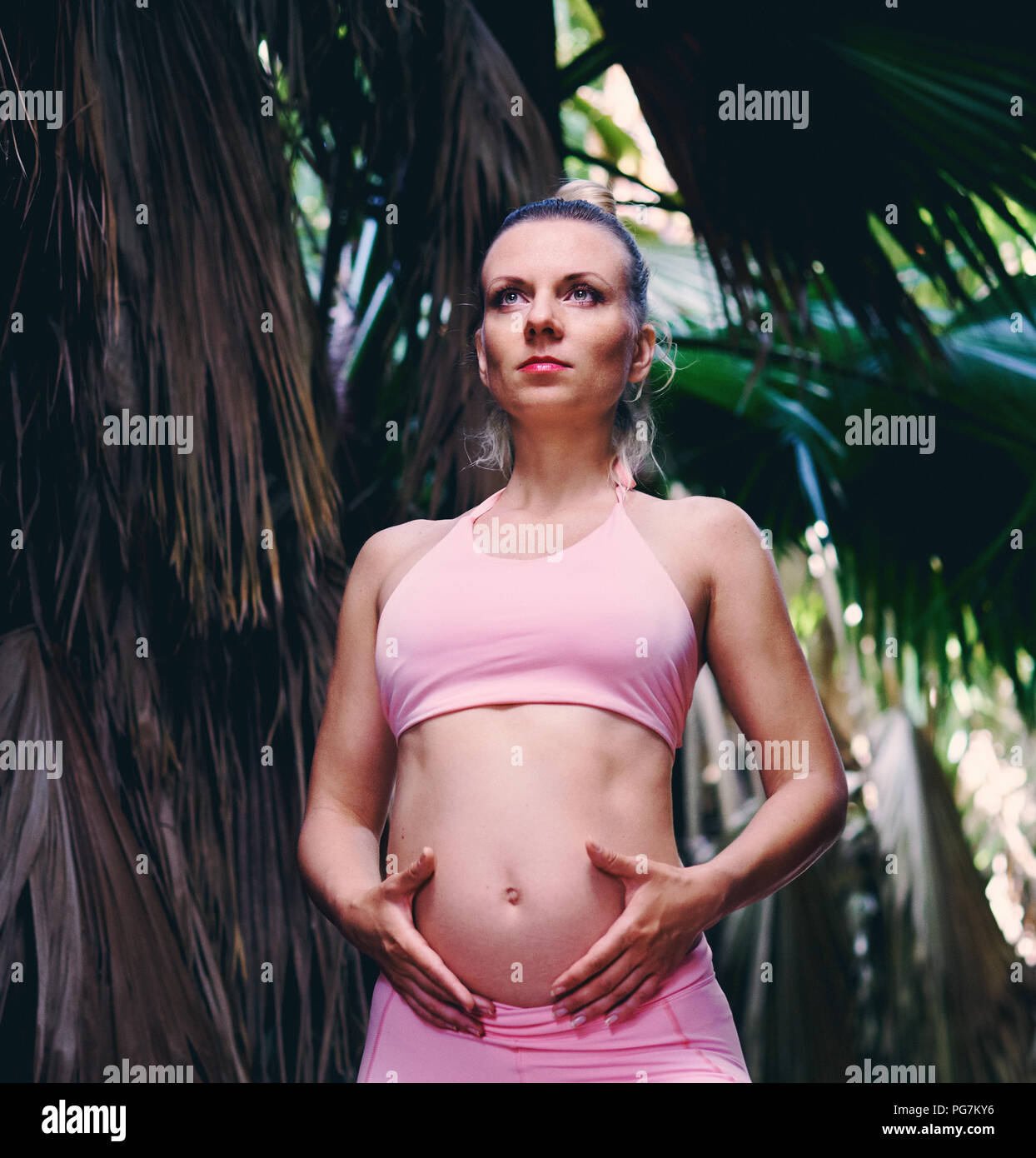 Young attractive blonde woman in sportswear rose faisant du yoga pilates exercices de fitness dans la nature tropicale, détient les mains sur le ventre. Avant la naissance de l'exercice, h Banque D'Images