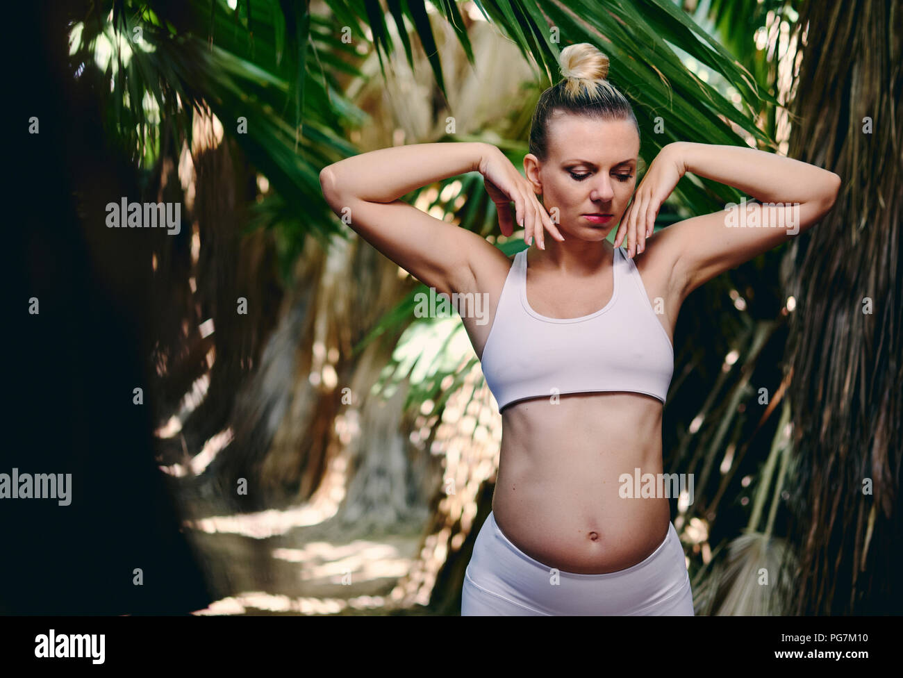 Young attractive blonde woman in white sportswear faisant du yoga pilates exercices de fitness dans la nature tropicale. Avant la naissance de l'exercice, mode de vie sain, rec Banque D'Images