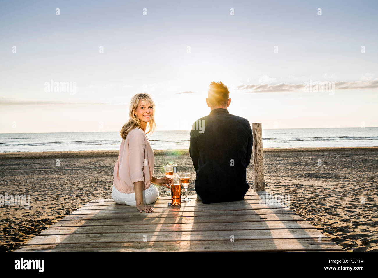 Couple avec des verres à vin assis sur la demande sur la plage au coucher du soleil Banque D'Images