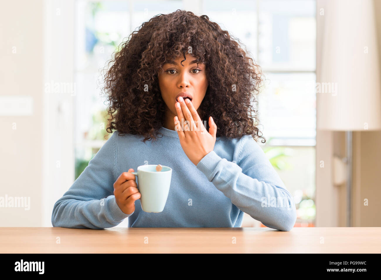 African American Woman holding une tasse de café à la maison couvrir la bouche à part choqué avec honte pour erreur, expression de la peur, peur en silence, se Banque D'Images