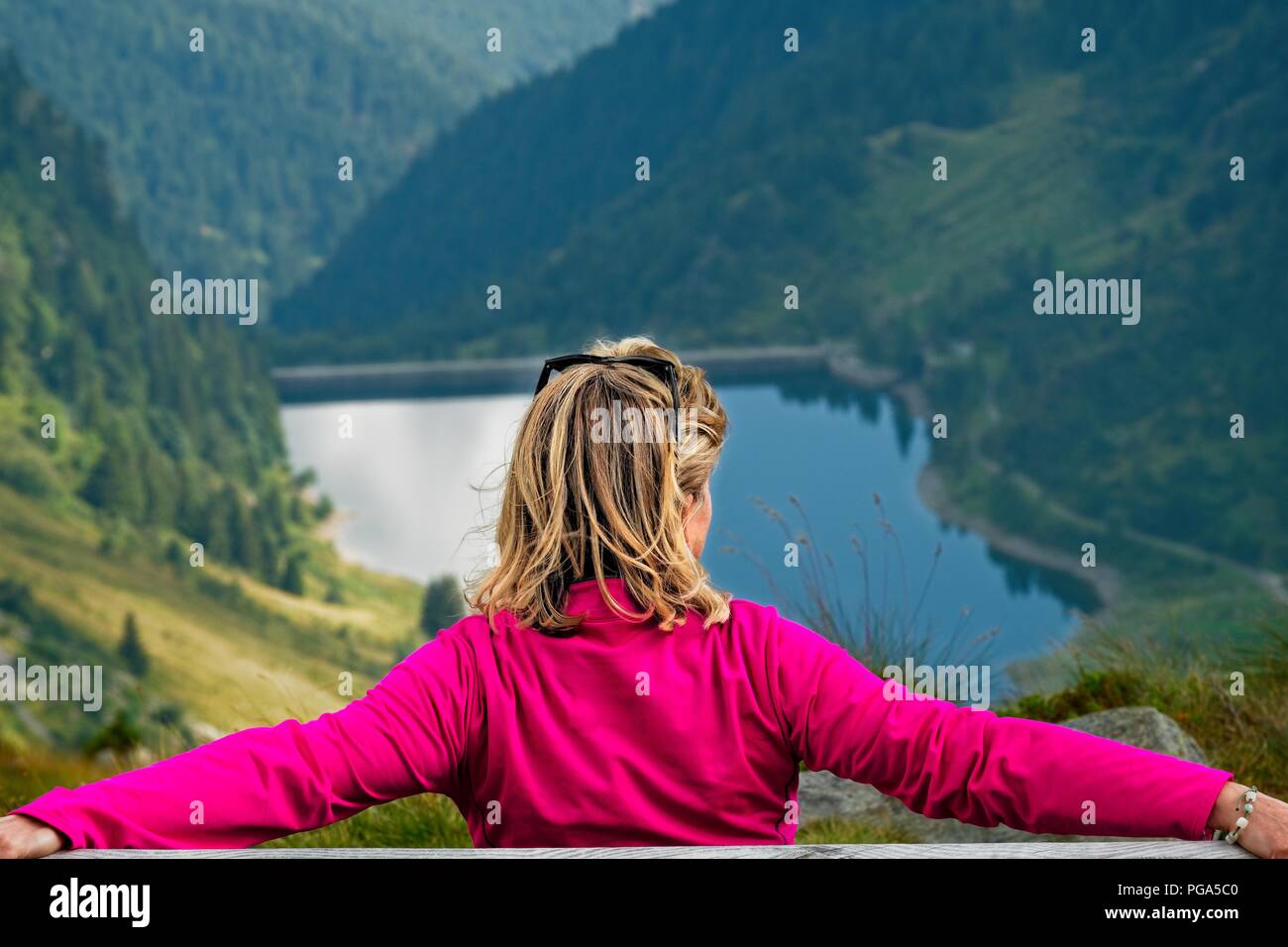 Une femme blonde assise sur un banc et à la recherche à l'horizon. Une vallée de montagne avec un lac est visible en arrière-plan. Tiré de l'arrière Banque D'Images