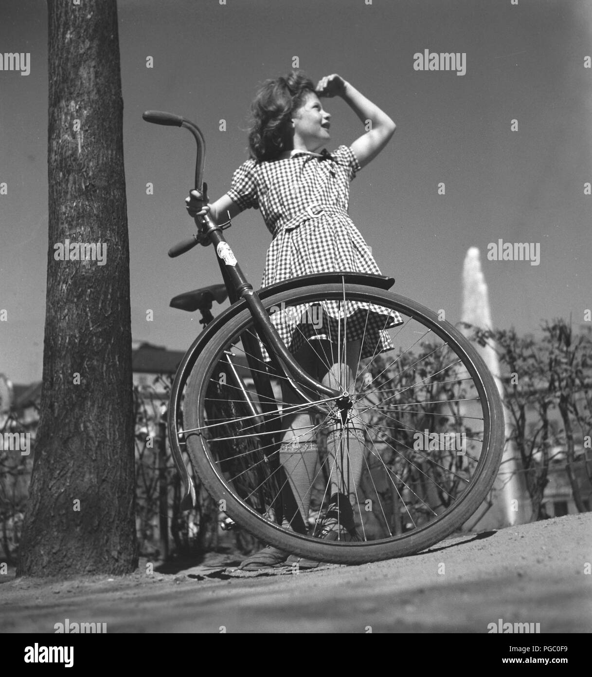 1940 fille avec location. Une jeune fille, habillée d'une robe à carreaux, est debout près de son vélo d'une journée ensoleillée. La Suède Mai 1940. 133-2 Kristoffersson Photo Banque D'Images