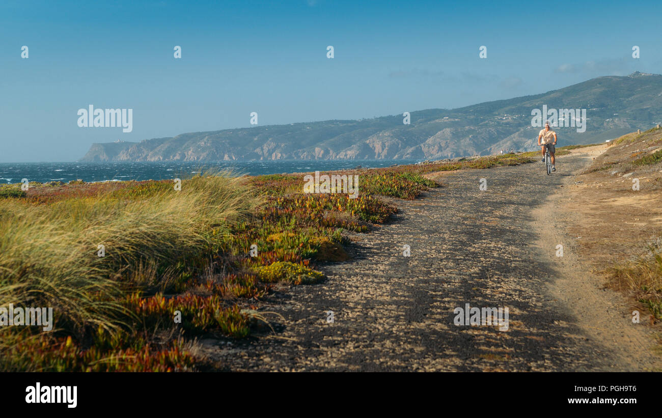 Homme mûr (55) cycles le long d'un chemin d'asphalte bosselée donnant sur Praia do Guincho et Roca Cabo près de Cascais, Portugal au cours d'une journée d'été venteux Banque D'Images
