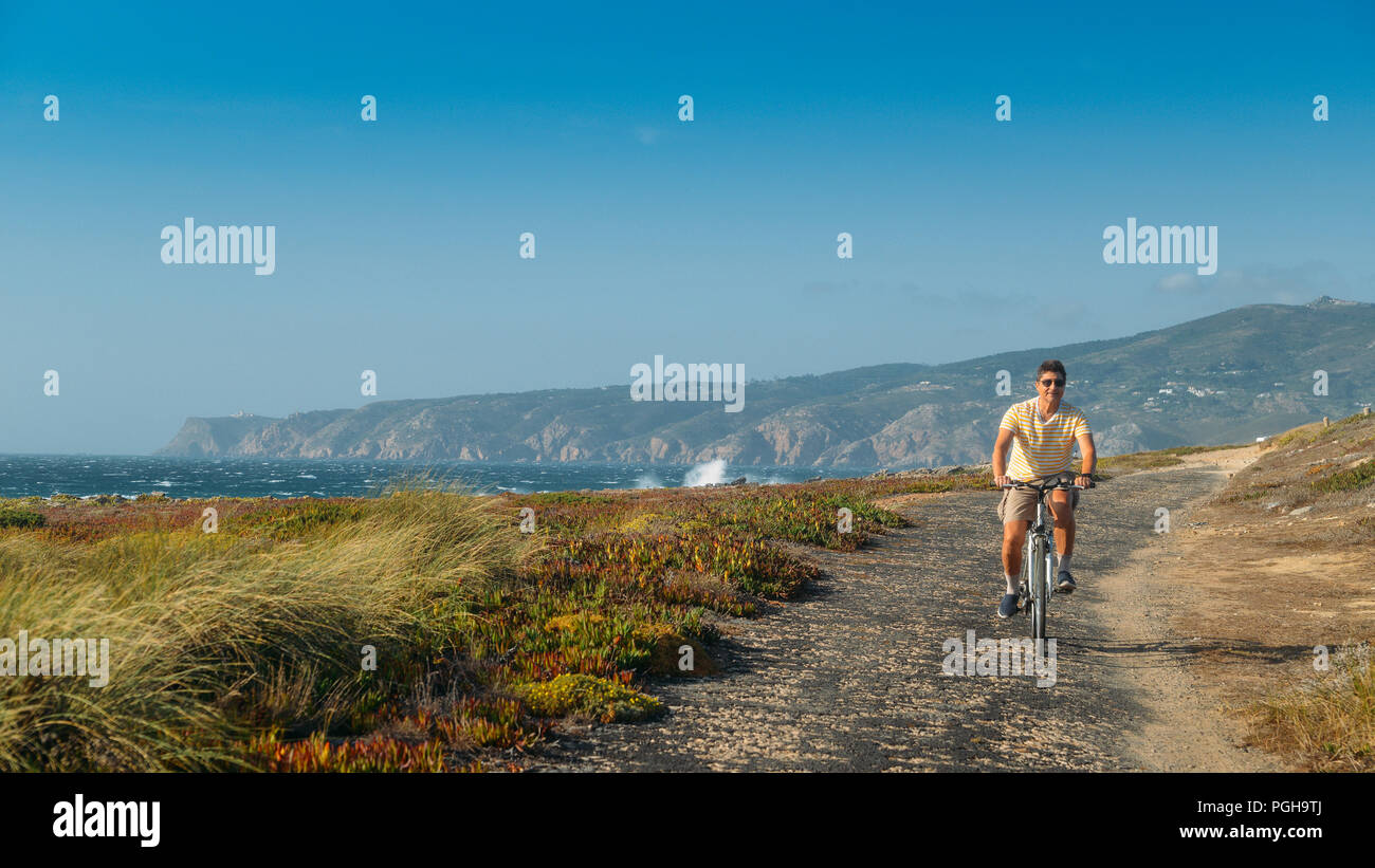 Homme mûr (55) cycles le long d'un chemin d'asphalte bosselée donnant sur Praia do Guincho et Roca Cabo près de Cascais, Portugal au cours d'une journée d'été venteux Banque D'Images
