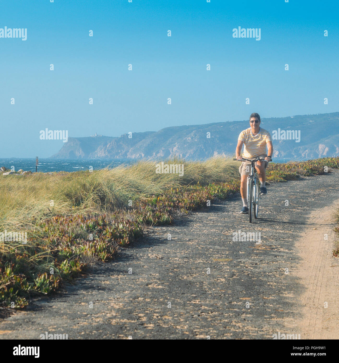 Homme mûr (55) cycles le long d'un chemin d'asphalte bosselée donnant sur Praia do Guincho et Roca Cabo près de Cascais, Portugal au cours d'une journée d'été venteux Banque D'Images