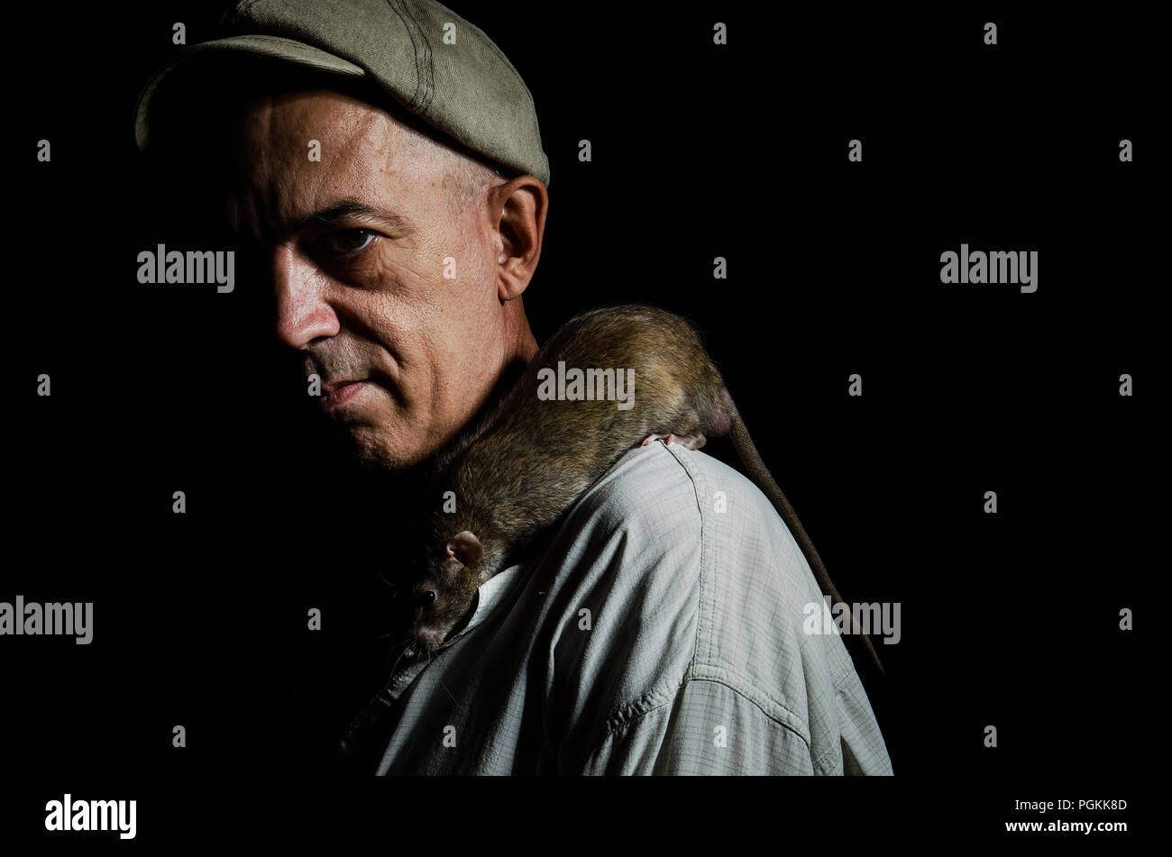 Un homme âgé dans un bouchon avec un rat domestique sur son épaule dans l'obscurité. Banque D'Images