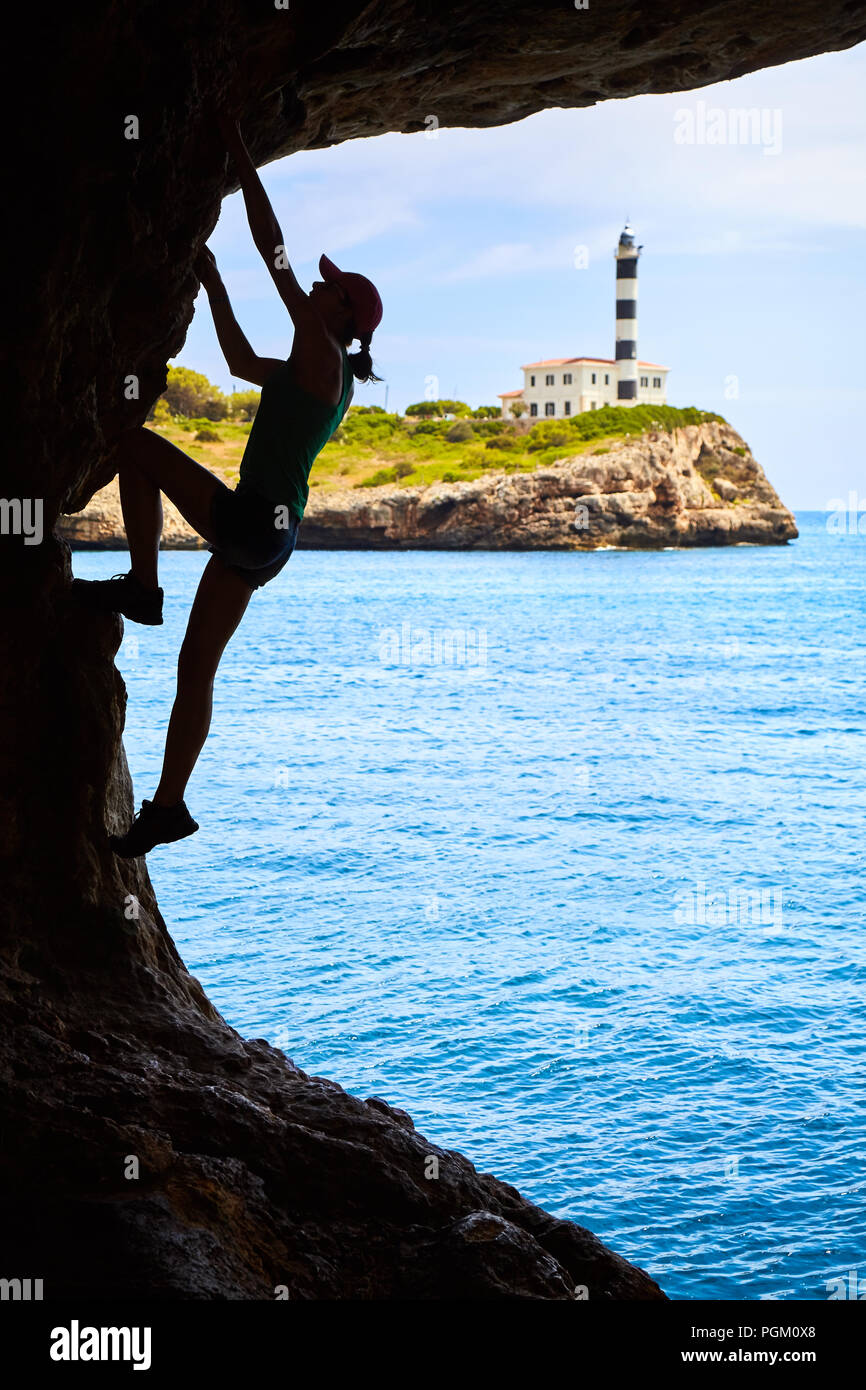 Athlète féminin de l'escalade dans une grotte, Portocolom Phare en arrière-plan, Mallorca, Espagne. Banque D'Images