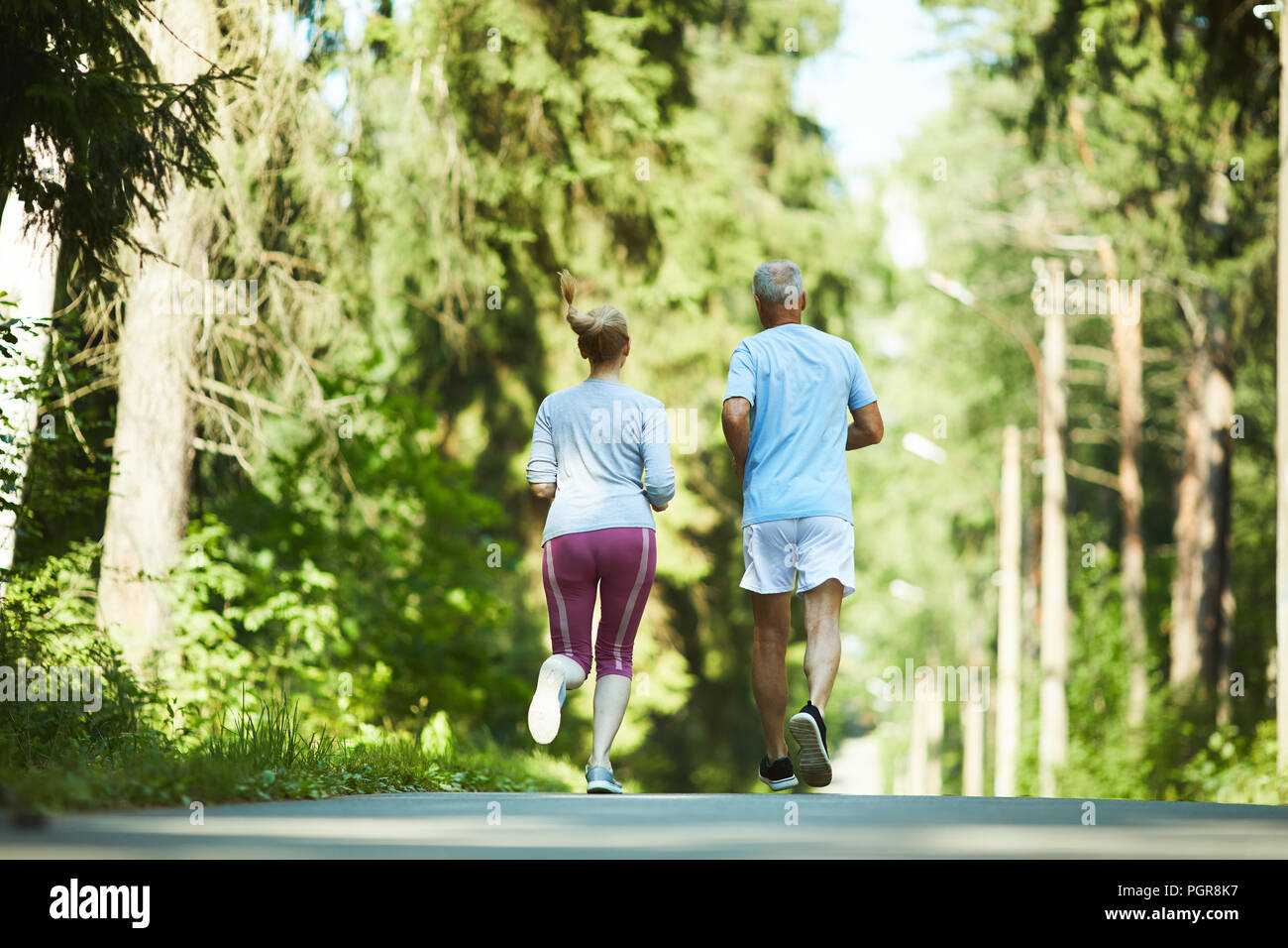 Vue arrière de l'âgés de conjoints dans activewear fonctionnant en bas de la route entre les arbres du parc sur sunny day Banque D'Images